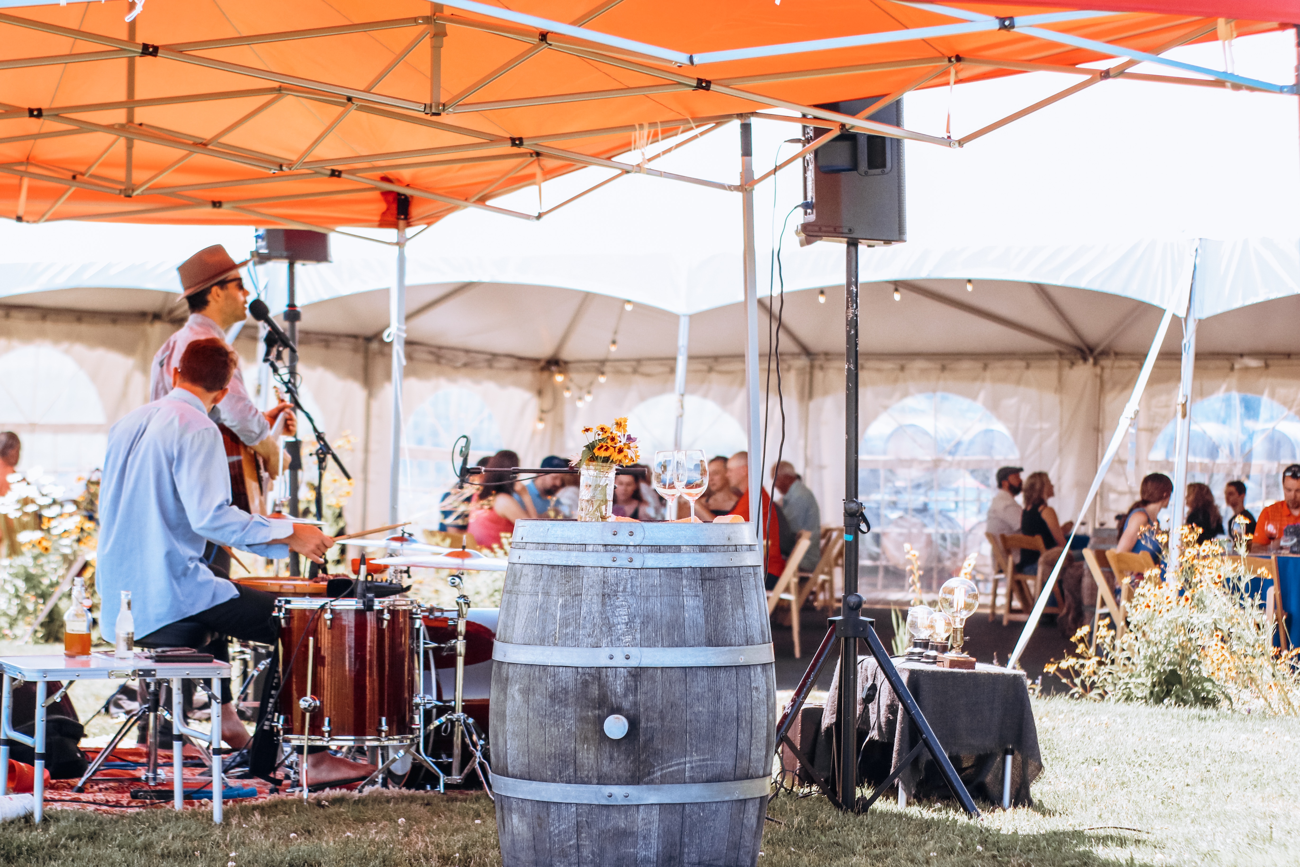 A concert takes place on the lawn at our Estate Tasting Room location under a large tent. In the foreground, a guitarist sings into a microphone while a drummer is mid-hit. In the background, several groups of people are seated at large banquet tables enjoying food and wine.